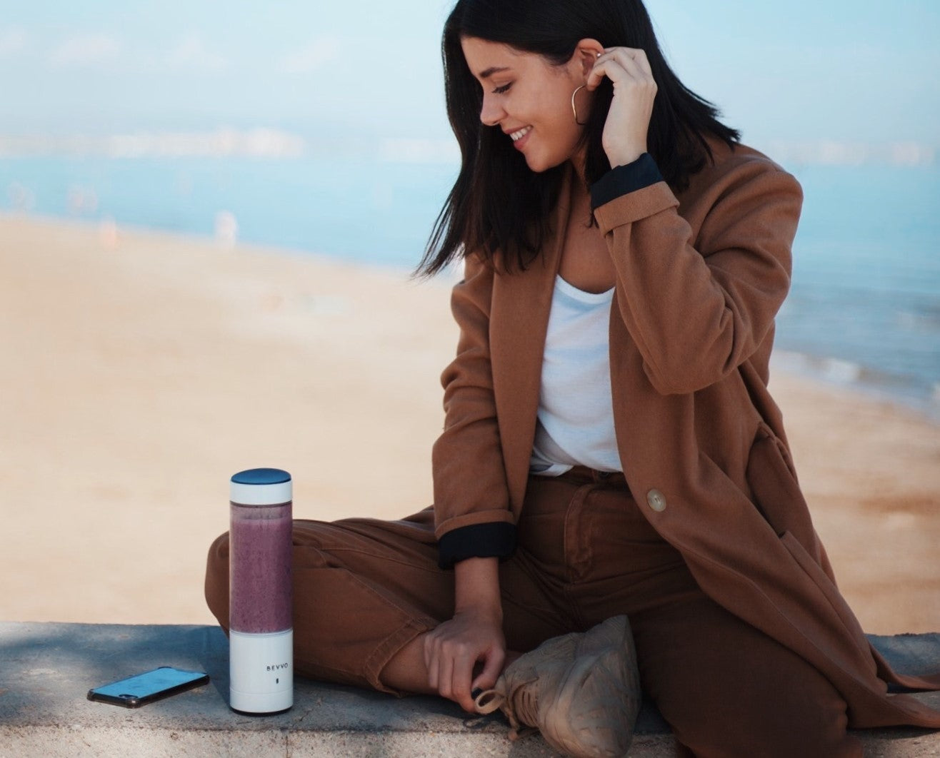 Woman sitting on ledge at the beach drinking smoothie from white BEVVO portable blender.